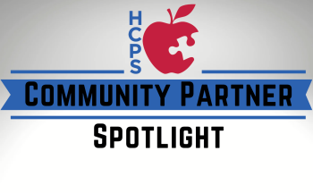 Logo for 'HCPS Community Partner Spotlight' featuring a red apple with a puzzle piece missing. The letters 'HCPS' are vertically aligned beside the apple, and the text 'Community Partner Spotlight' is displayed prominently across the center with blue and black accents.