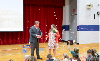 Ms. Ashley McVey and Superintendent Mark Garrett stand in front of the gym with confetti during the announcement. There are students sitting on the gym floor.