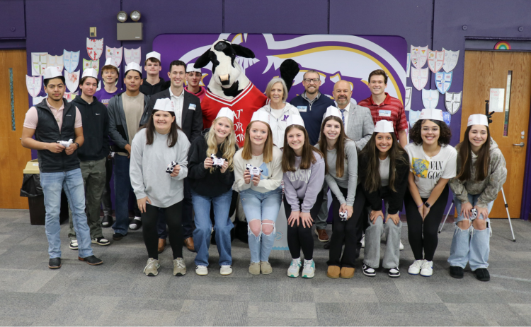 14 leadership students, Coach Sue Moon, and four Chick-fil-a members pose for a photo with the Chick-fil-a cow. The students are wearing Chick-fil-a paper hats and holding mini cows. The backdrop is a North Henderson Knight logo in purple. The location is the school media center