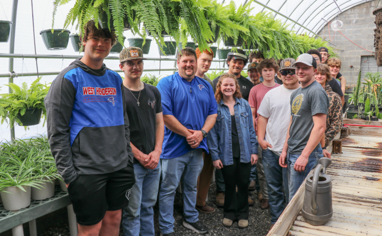 Jason Livingston poses with several students in the school green house for a group photo. There are several plants and ferns in the background